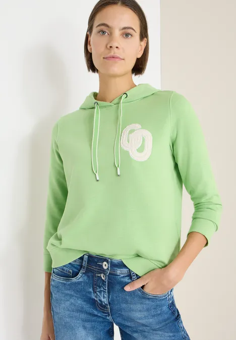 Frau trägt hellgrünen Hoodie und Jeans, eine Hand in Tasche, lässiger Stil.